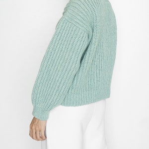 May include: A light sage green knit sweater featuring a chunky ribbed texture, a crew neckline, and long sleeves with fitted cuffs. The garment has a relaxed fit and a thick, woven construction that provides a distinct, tactile appearance.