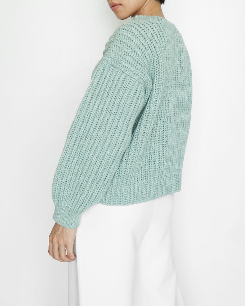 May include: A light sage green knit sweater featuring a chunky ribbed texture, a crew neckline, and long sleeves with fitted cuffs. The garment has a relaxed fit and a thick, woven construction that provides a distinct, tactile appearance.