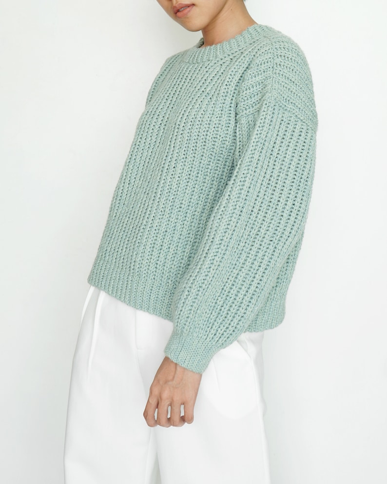 May include: A light sage green long-sleeve sweater featuring a chunky ribbed knit texture, a crew neckline, and dropped shoulders. The garment is paired with plain white trousers, creating a clean and simple look with a relaxed fit.