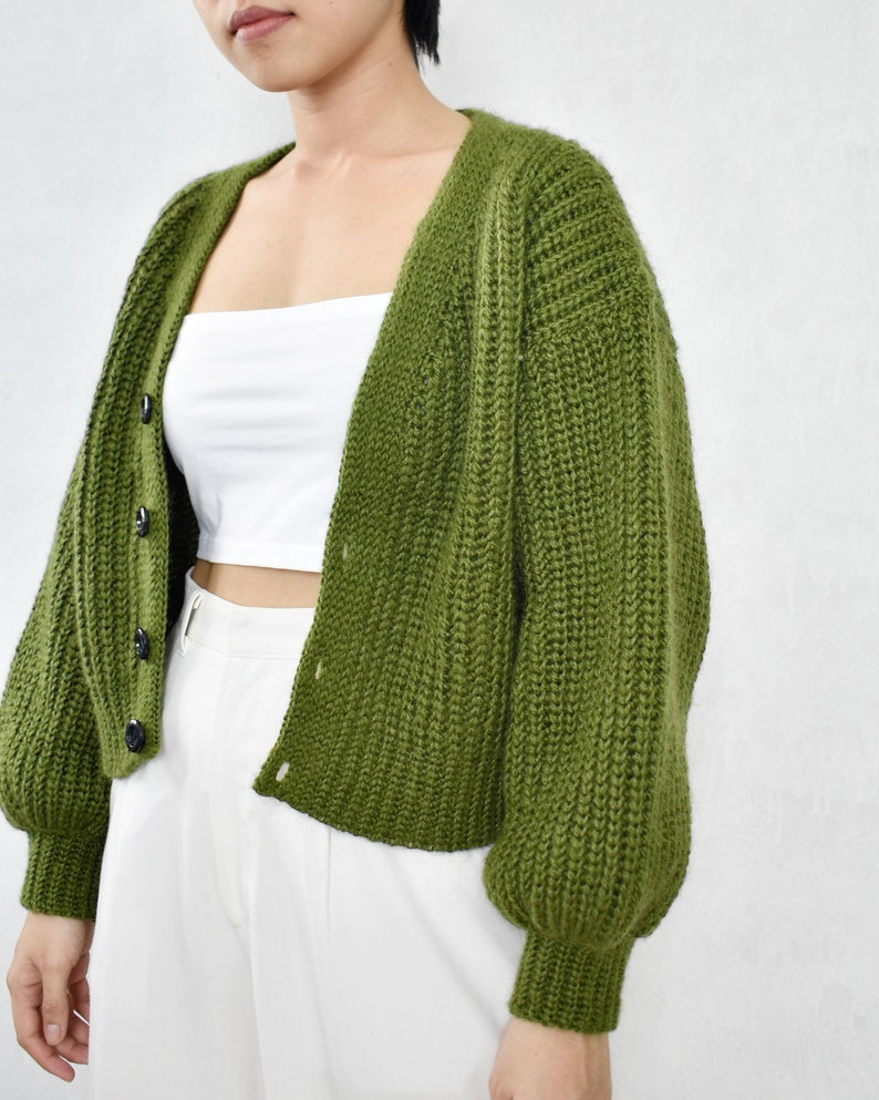 May include: A green knitted cardigan with a V-neck and button fastening. The cardigan has long sleeves with puffed shoulders and a cropped length.