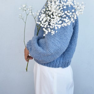 May include: A light blue knitted sweater with long sleeves and a relaxed fit. The sweater is worn by a person holding a bouquet of white baby's breath flowers.