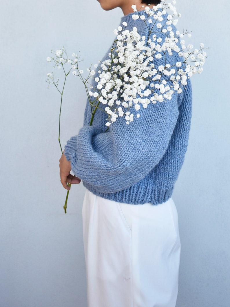 May include: A light blue knitted sweater with long sleeves and a relaxed fit. The sweater is worn by a person holding a bouquet of white baby's breath flowers.