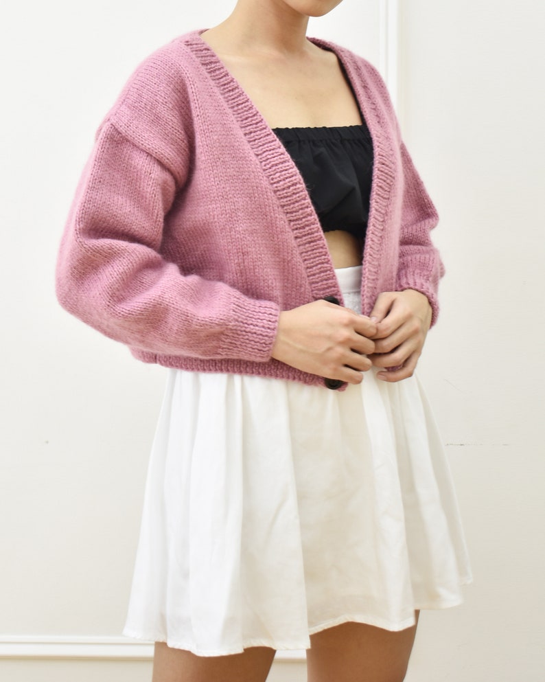 May include: A pink knitted cardigan with a single button closure. The cardigan has a V-neck and long sleeves.