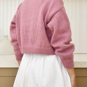 May include: A pink knitted cropped sweater with long sleeves. The sweater is made of a soft, fuzzy yarn.