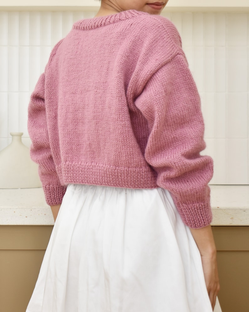 May include: A pink knitted cropped sweater with long sleeves. The sweater is made of a soft, fuzzy yarn.