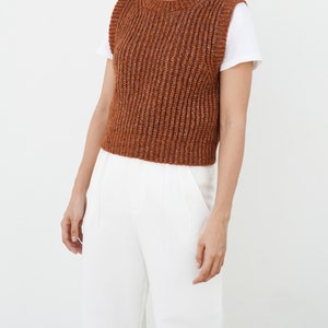 K&ouml;nnte beinhalten: Eine braune, gestrickte, &auml;rmellose Strickweste mit Rippenstruktur. Die Weste wird &uuml;ber einem wei&szlig;en T-Shirt und einer wei&szlig;en Hose getragen.