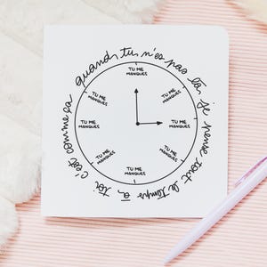 Puede incluir: Una tarjeta cuadrada blanca con un diseño de reloj y texto en francés, junto con un bolígrafo blanco. La esfera del reloj dice "Tu me manques" (Te echo de menos) a cada hora. La tarjeta está sobre una superficie rosa.