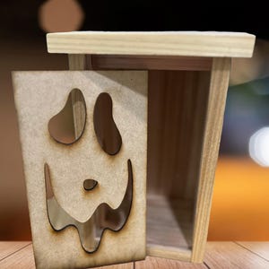 May include: A wooden box with a cutout face design. The box is made of light-colored wood and has a rectangular shape with a lid. The face design features large eye shapes and a curved mouth, creating a smiling expression. The box is likely a decorative item.