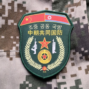 以下が含まれることがあります： 朝鮮語で「朝鮮共同国防」と中国語で「中華人民共和国国防」と書かれた緑色の軍用パッチ。パッチには赤い星、2丁のライフル、葉の輪が描かれている。