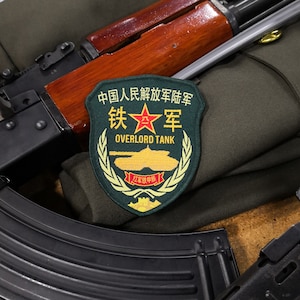 May include: A close-up of a military patch with a red star and the text "中国人民解放军铁军 OVERLORD TANK" in Chinese and English. The patch is on a green fabric background with a black and brown rifle and a black magazine.