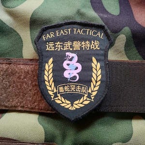 May include: A black and gold patch with the text "FAR EAST TACTICAL" and Chinese characters. The patch features a snake and lightning bolt design, surrounded by a gold wreath, on a camouflage background.