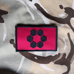 May include: A red embroidered patch with a black hexagonal design. The patch has a black border and is sewn onto a camouflage fabric.