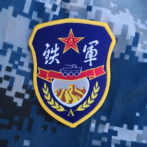 Peut inclure: Écusson en forme de bouclier avec une bordure jaune, des caractères chinois et une étoile rouge en haut. En dessous, une illustration de char et un paysage. L'écusson est posé sur un fond de camouflage bleu et gris.