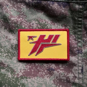 May include: A red and yellow embroidered patch with a stylized airplane and the letter "H" in a red font. The patch is sewn onto a camouflage fabric.