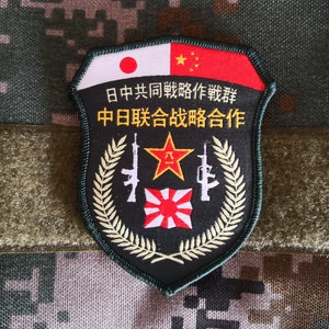 May include: A black and green military patch with a red star, two rifles, and a wreath of leaves. The patch has the text "日中共同戦勝作戦" and "中日联合战勝合作" in Japanese and Chinese.