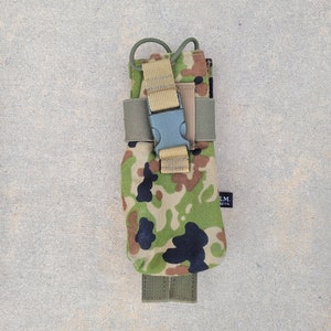 以下が含まれることがあります： 迷彩柄のポーチで、タンのストラップと黒いバックルが付いています。ポーチの下部に緑色のストラップが付いており、側面には「E.M. INC.」の文字が印刷されています。