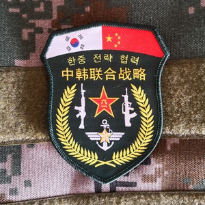 May include: A military patch with the flags of South Korea and China at the top. The patch is green with gold accents, a red star, rifles, and text in Korean and Chinese. The background is a camouflage pattern.