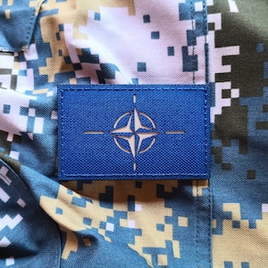 Può includere: Una toppa blu con una stella bianca e quattro linee bianche che formano una forma di bussola. La toppa è su uno sfondo di tessuto mimetico.