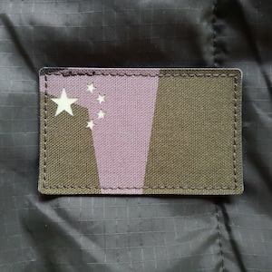 Può includere: Una toppa di mimetica marrone e rosa con cinque stelle bianche. La toppa è cucita con filo bianco.