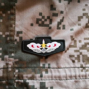 Puede incluir: Un parche militar negro con una estrella blanca, un diseño de nube roja y blanca y un misil. El parche está sobre una tela de camuflaje.
