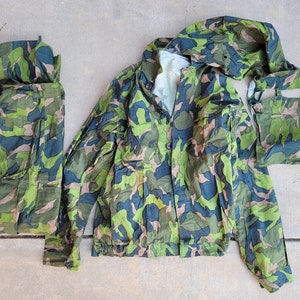 May include: Camouflage military jacket, pants, and hood. The jacket has a button closure and a hood. The pants have a zipper and button closure. The hood is a separate piece.