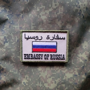 Könnte beinhalten: Ein beigefarbener gestickter Aufnäher mit der russischen Flagge und dem Text "Embassy of Russia" in Schwarz. Der Text "سفارة روسيا" ist in arabischer Schrift über der Flagge geschrieben.