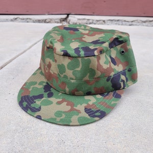 以下が含まれることがあります： 緑、茶色、黒の迷彩柄の軍用スタイルの帽子。帽子は硬いつばと上部にボタンが付いています。