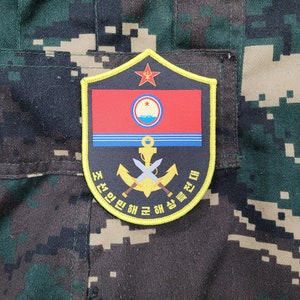 May include: A yellow and black embroidered patch with a red and blue flag, a crossed anchor and swords, and the text "조선 민주주의 인민 공화국 해군" in Korean.