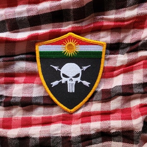 Può includere: Una toppa ricamata nera e gialla con un teschio e ossa incrociate, un sole giallo e strisce verdi e bianche. La toppa è su un tessuto a quadri rossi e bianchi.