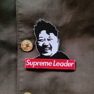 Può includere: Una toppa in bianco e nero del viso di un uomo con il testo "Supreme Leader" in lettere bianche su sfondo rosso.