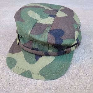 Könnte beinhalten: Eine Militärkappe im Tarnmuster in Grün mit einem schwarzen verstellbaren Riemen.