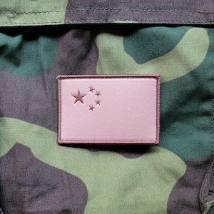 Può includere: Un cerotto di stoffa rosa con la bandiera della Cina. La bandiera presenta cinque stelle gialle, una grande e quattro più piccole, su un campo rosso. Il cerotto è cucito su un tessuto mimetico verde, marrone e nero.