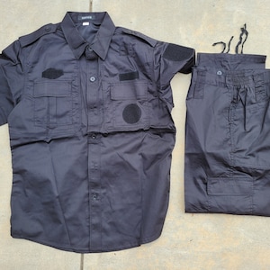 Peut inclure: Une chemise noire à manches courtes et un pantalon assorti avec plusieurs poches et des patchs en velcro. La chemise a un devant boutonné et un col.
