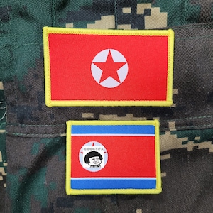 Op de afbeelding: Twee rechthoekige patches met het Noord-Koreaanse vlagontwerp. De bovenste patch heeft een rode achtergrond met een witte ster in het midden, omlijnd in geel. De onderste patch heeft een rode achtergrond met een cartoonafbeelding en tekst.