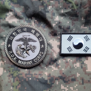 Puede incluir: Un parche bordado gris y negro con el texto "R.O.K. MARINE CORPS" y un águila estilizada, un ancla y una estrella. El parche está sobre un fondo de tela de camuflaje. Un parche de la bandera surcoreana blanco y negro también está sobre la tela.