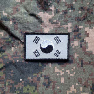 以下が含まれることがあります： 韓国の国旗の白黒刺繍パッチ。旗は白い背景に中央に黒い円があります。円の中に白い陰陽のシンボルがあります。旗は黒い縁取りで囲まれています。