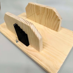 Può includere: Una guida per tagliare il sapone in legno con un tenditore di filo in plastica nera. La guida ha due fessure verticali per tenere le barre di sapone e un filo che attraversa la parte superiore per tagliare il sapone.