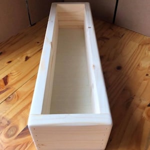 Puede incluir: Una caja de madera larga y rectangular con un acabado natural. La caja está vacía y tiene una superficie lisa.