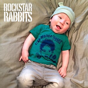 Op de afbeelding: Een baby die een groen T-shirt draagt met een blauwe afbeelding waarop staat "My Mother wears", een grijze beanie en een grijze spijkerbroek met witte sneakers.