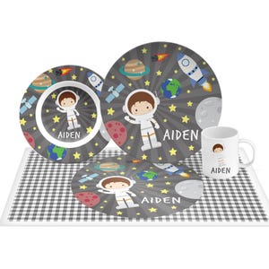 May include: A dinnerware set featuring a bowl, a plate, a mug, and a placemat, all decorated with a space theme including planets, rockets, stars, and an astronaut character. Each piece is personalized with the name AIDEN in white text.