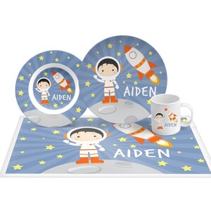 May include: A dinnerware set featuring a plate, bowl, and mug with a blue space theme, yellow stars, a rocket, and an astronaut character on the moon. Each piece is personalized with the name AIDEN in white text against the blue background.