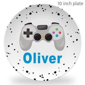 May include: A 10 inch round plate with a white surface featuring black speckles, a gray video game controller illustration, and the name Oliver printed in blue text. The design includes a black directional pad and four colored buttons.