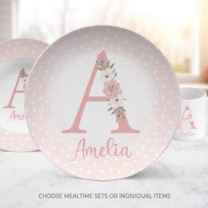 May include: A light pink plate and mug set featuring a white polka dot pattern. The plate displays a large pink letter A decorated with a floral arrangement of pink and white flowers, with the name Amelia written in pink script below the letter.