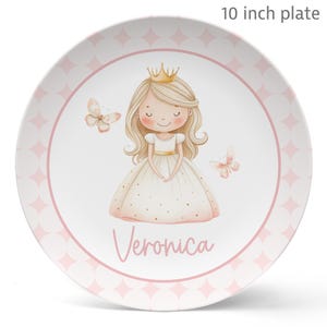 May include: A 10-inch plate featuring a princess illustration. The design includes a blonde princess wearing a gold crown and a white dress with gold dots. Two butterflies and the name "Veronica" are also featured, all set against a pink and white background.