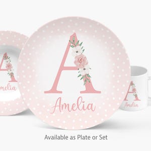 May include: A pink polka-dotted plate with a large pink letter "A" decorated with flowers and leaves. The name "Amelia" is written below the letter in cursive. A matching plate and mug are also visible.