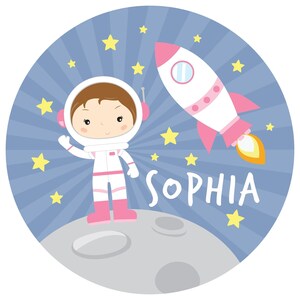 May include: A circular graphic featuring a cartoon astronaut in a white and pink suit standing on a gray moon surface. A white and pink rocket ship with a yellow flame flies in the blue background with yellow stars. The name SOPHIA is printed in white.