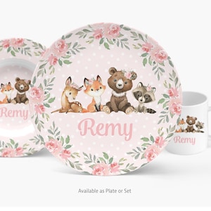 May include: A pink plate with a floral border and a woodland animal design. The plate features a deer, fox, bear, and raccoon. The name "Remy" is written in pink script. A matching mug and bowl are also visible.