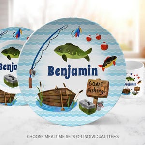 May include: A round plate featuring a light blue wavy water pattern, a green fish, a fishing rod, a wooden boat, a tackle box, and a sign that reads Gone Fishing. The name Benjamin is printed in dark blue text in the center of the plate.