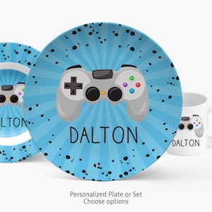 May include: A round plate and a white mug featuring a light blue sunburst background with black paint splatters. A gray video game controller graphic is centered on each item, with the name DALTON printed in black capital letters below it.