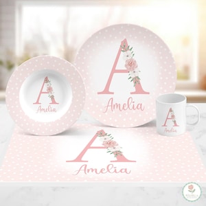 May include: A dinnerware set featuring a plate, bowl, mug, and placemat, all in light pink with white polka dots. Each piece displays a large pink letter A decorated with watercolor flowers and the name Amelia printed in pink script lettering.
